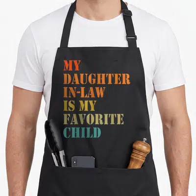 Retro daughter father day apron - Stacked colorful text reading MY DAUGHTER IN-LAW IS MY FAVORITE CHILD in red orange — shown on a black cotton canvas apron against a white background