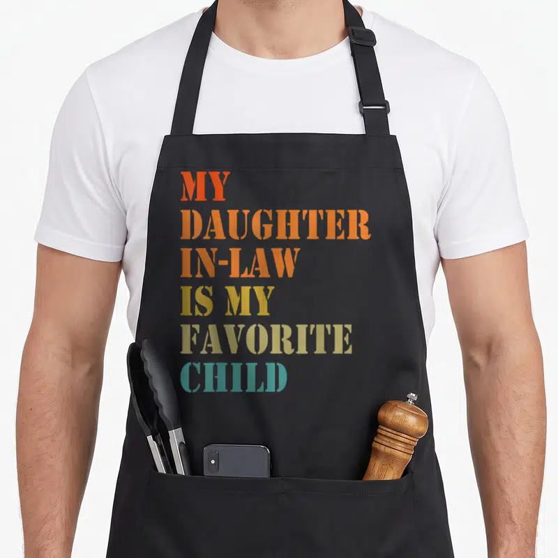 Retro daughter father day apron - Stacked colorful text reading MY DAUGHTER IN-LAW IS MY FAVORITE CHILD in red orange — shown on a black cotton canvas apron against a white background