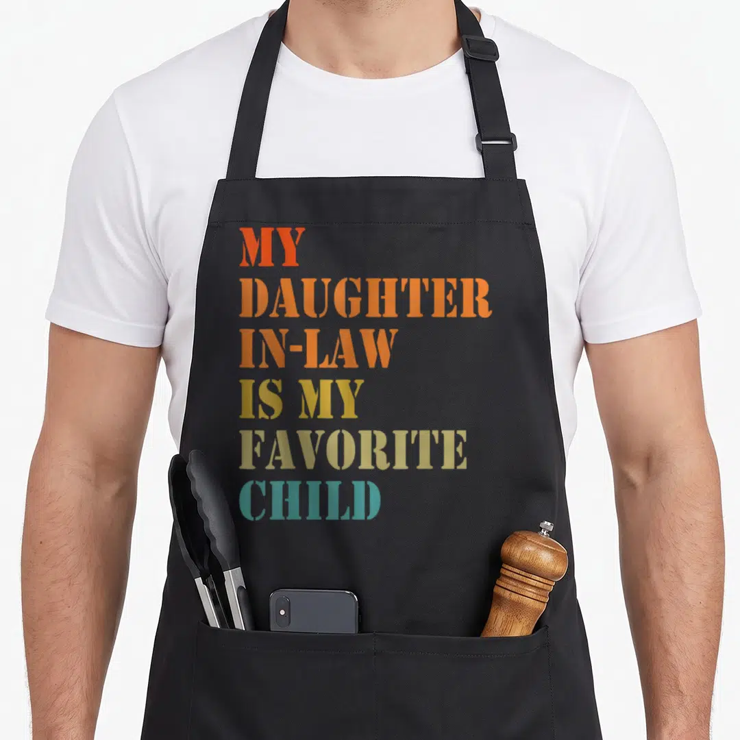 Retro daughter father day apron - Stacked colorful text reading MY DAUGHTER IN-LAW IS MY FAVORITE CHILD in red orange — shown on a black cotton canvas apron against a white background