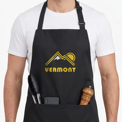 Funny mountain vermont apron - Yellow distressed mountain range graphic above white peaks and bold VERMONT text — shown on a black cotton canvas apron against a white background