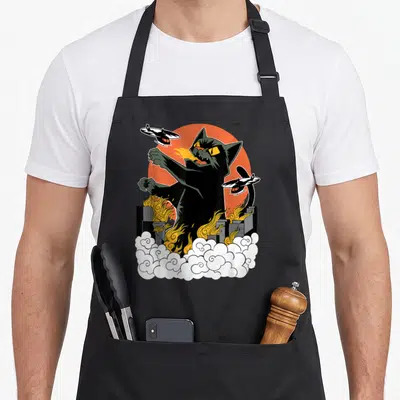 Japanese crew apron - Black cat breathing fire over city with helicopters and orange sun cartoon — shown on a black cotton canvas apron against a white background
