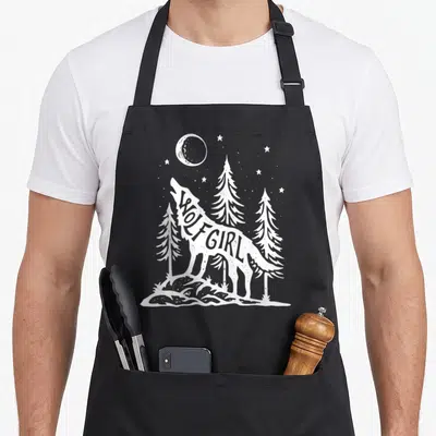 Wolf outdoor apron - White wolf silhouette containing WOLF GIRL text with pine trees and moon — shown on a black cotton canvas apron against a white background