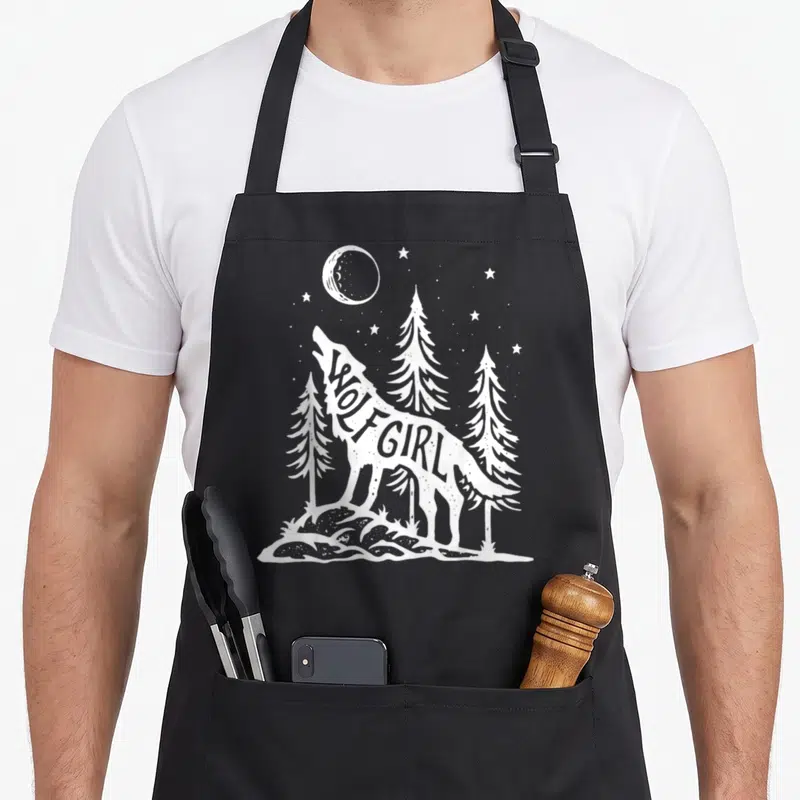 Wolf outdoor apron - White wolf silhouette containing WOLF GIRL text with pine trees and moon — shown on a black cotton canvas apron against a white background