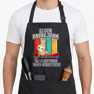 Canoeing sloth apron - Cartoon brown sloth in red kayak with SLOTH KAYAK TEAM text on multi-colored striped background — shown on a black cotton canvas apron against a white background