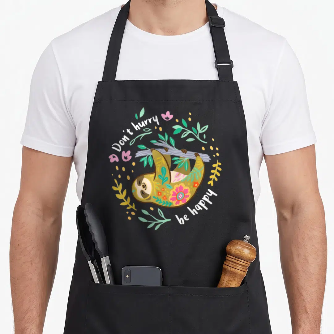 Hurry sloth apron - Cartoon sloth hanging from grey branch with floral circle and text Don't hurry be happy — shown on a black cotton canvas apron against a white background