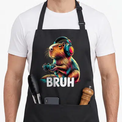 Capybaras video game apron - Cartoon capybara with headphones and controller holding BRUH text in brown blue pink — shown on a black cotton canvas apron against a white background