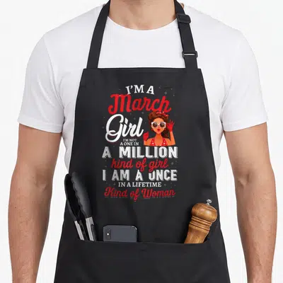 Retro march birthday apron - Cartoon woman with sunglasses and red text reading I am a once in a lifetime kind of woman — shown on a black cotton canvas apron against a white background