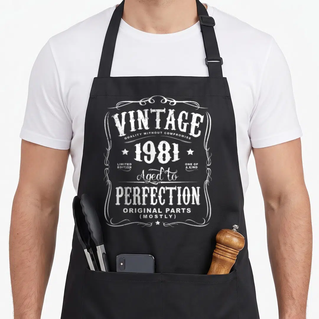 37th birthday apron - White distressed vintage 1981 text design with ornate frame and stars — shown on a black cotton canvas apron against a white background