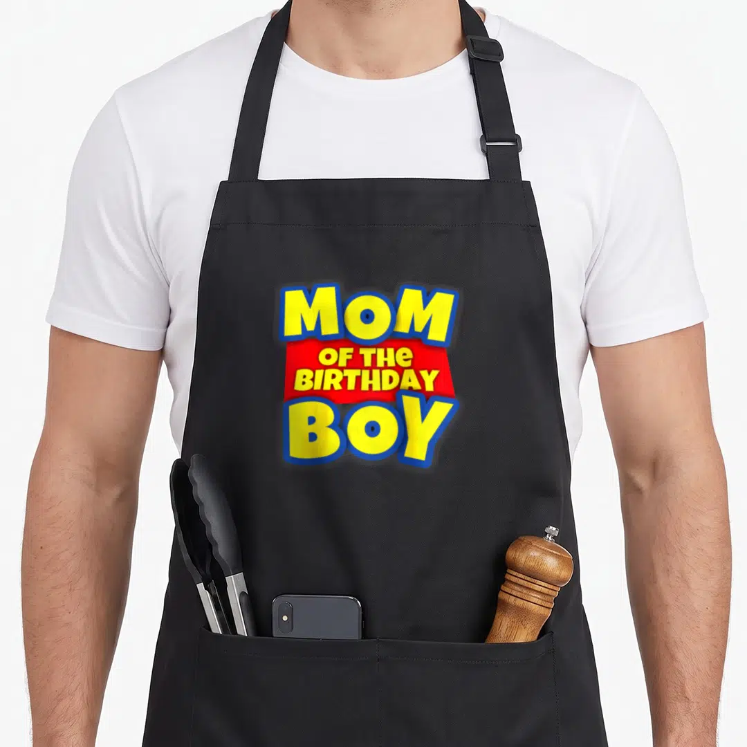 Toy baseball birthday apron - MOM OF THE BIRTHDAY BOY text in yellow blue and red stacked typography — shown on a black cotton canvas apron against a white background