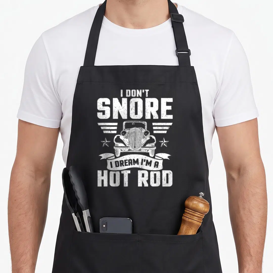 Snore hot rod apron - White distressed hot rod illustration with stars and ribbon text reading I Don't Snore I Dream — shown on a black cotton canvas apron against a white background