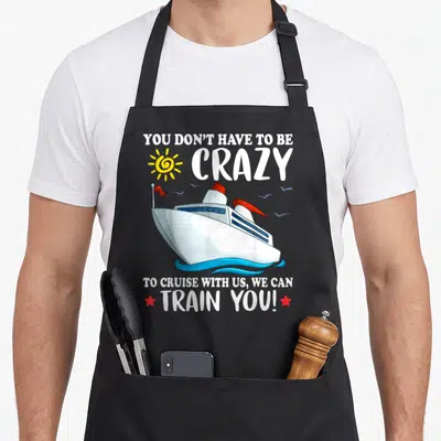 Cruise train us aprons - Cartoon cruise ship on waves with pun text YOU DON'T HAVE TO BE CRAZY TO CRUISE WITH US WE CAN — shown on a black cotton canvas apron against a white background