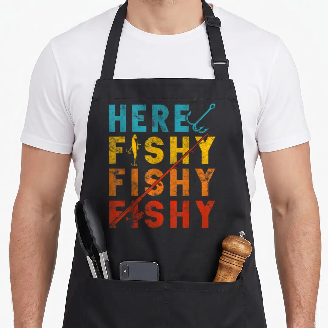 Fisher fishing bait apron - Stacked HERE FISHY FISHY FISHY text in blue yellow orange red with red rod and blue hook — shown on a black cotton canvas apron against a white background