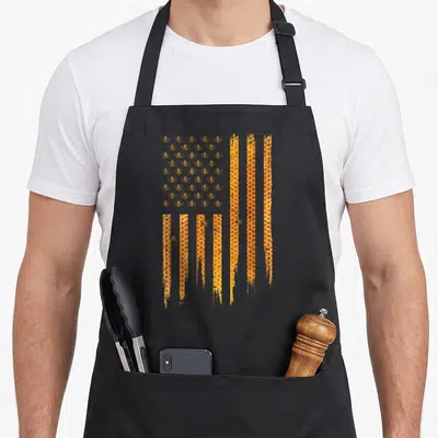 Honey american flag apron - USA flag pattern made of bees and vertical honeycomb columns in orange and yellow — shown on a black cotton canvas apron against a white background
