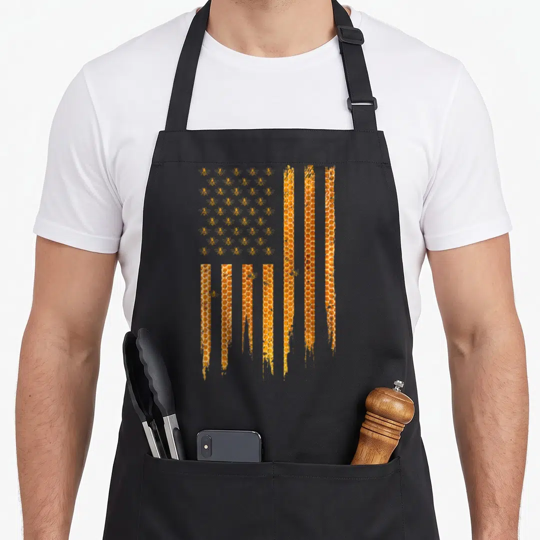 Honey american flag apron - USA flag pattern made of bees and vertical honeycomb columns in orange and yellow — shown on a black cotton canvas apron against a white background