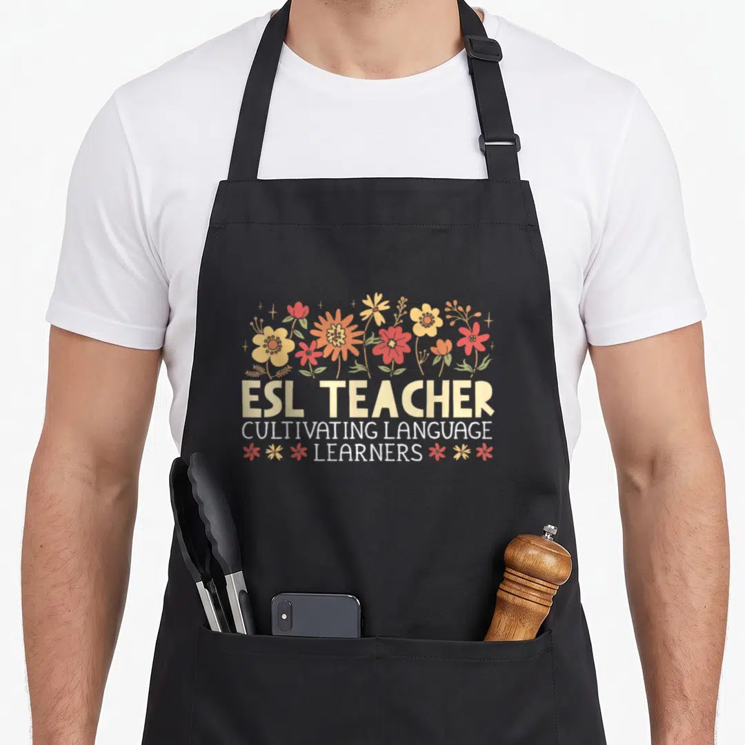 Esl english chef apron - ESL TEACHER text surrounded by cartoon flowers in orange, red, and yellow with green leaves — shown on a black cotton canvas apron against a white background