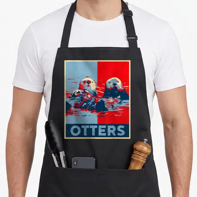 Sea otter man apron - Two otters on split blue red background with OTTERS text in blue bar below — shown on a black cotton canvas apron against a white background