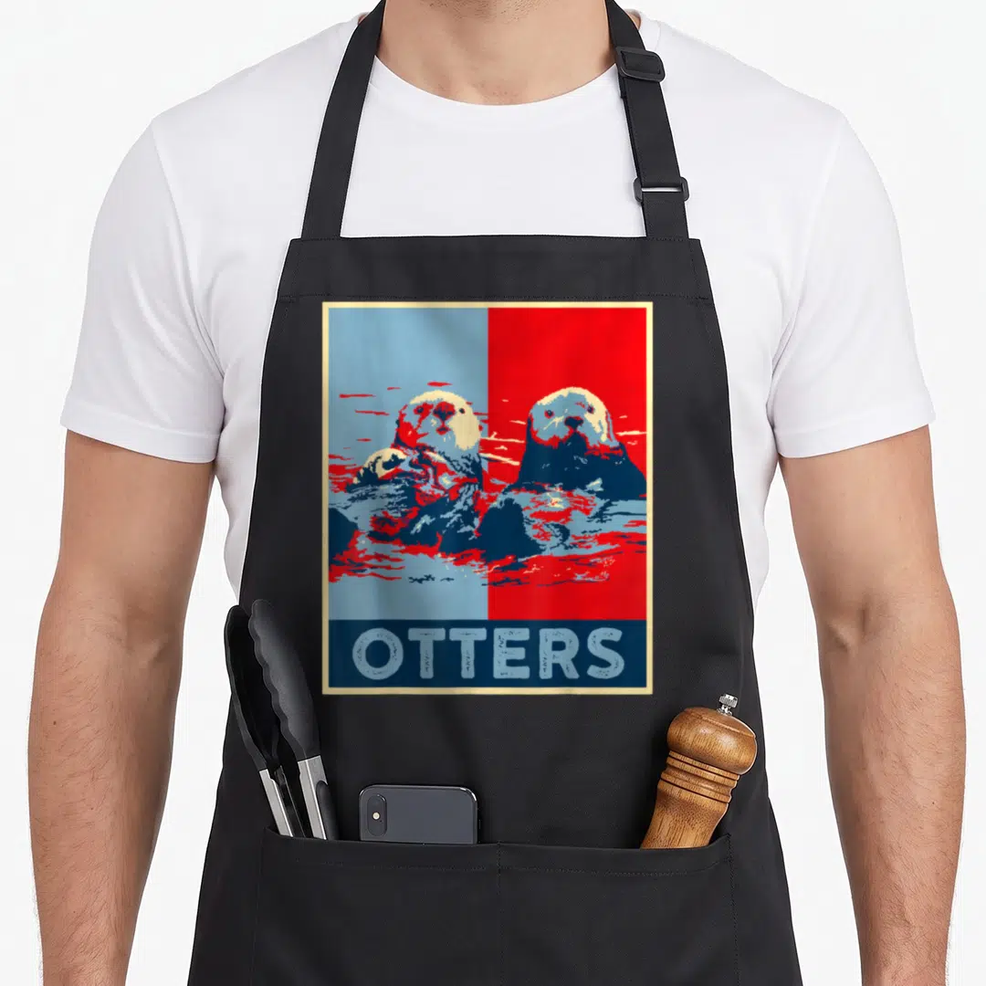 Sea otter man apron - Two otters on split blue red background with OTTERS text in blue bar below — shown on a black cotton canvas apron against a white background