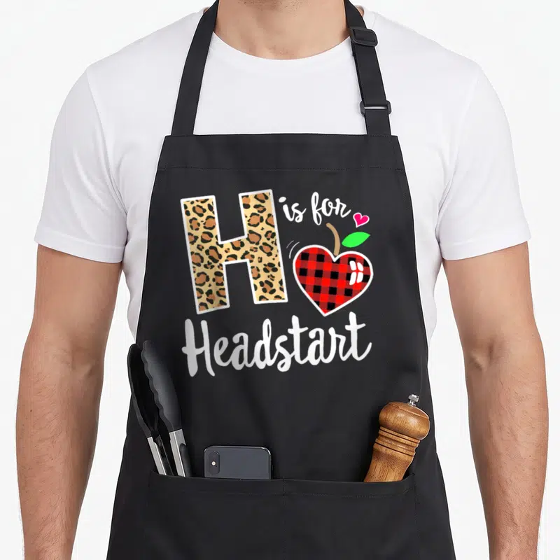 Headstart the plaid apron - Leopard print letter H next to red black plaid apple and white script text — shown on a black cotton canvas apron against a white background