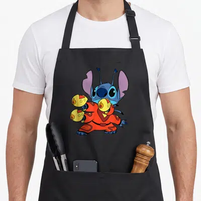 Space fighter apron - Blue cartoon character in orange suit holding yellow spheres with purple ears standing — shown on a black cotton canvas apron against a white background