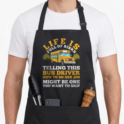 Bus driving apron - Cartoon yellow school bus between two trees with sarcastic text about bus driver risks — shown on a black cotton canvas apron against a white background