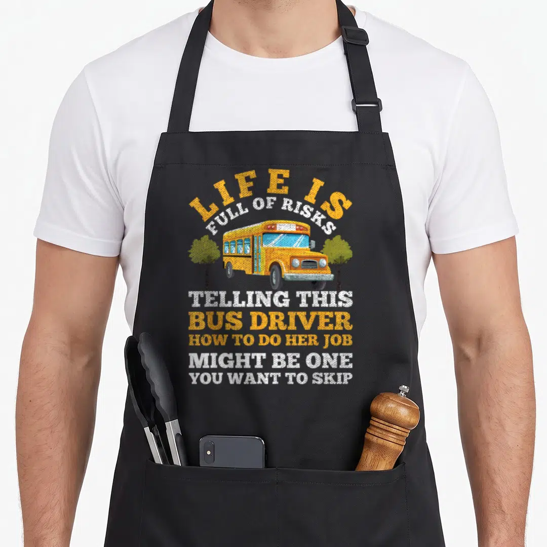 Bus driving apron - Cartoon yellow school bus between two trees with sarcastic text about bus driver risks — shown on a black cotton canvas apron against a white background