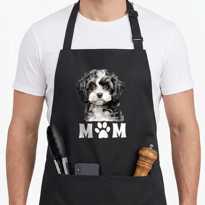 Mum dog face apron - Realistic tricolor dog portrait above white MOM text with a paw print replacing the middle letter — shown on a black cotton canvas apron against a white background