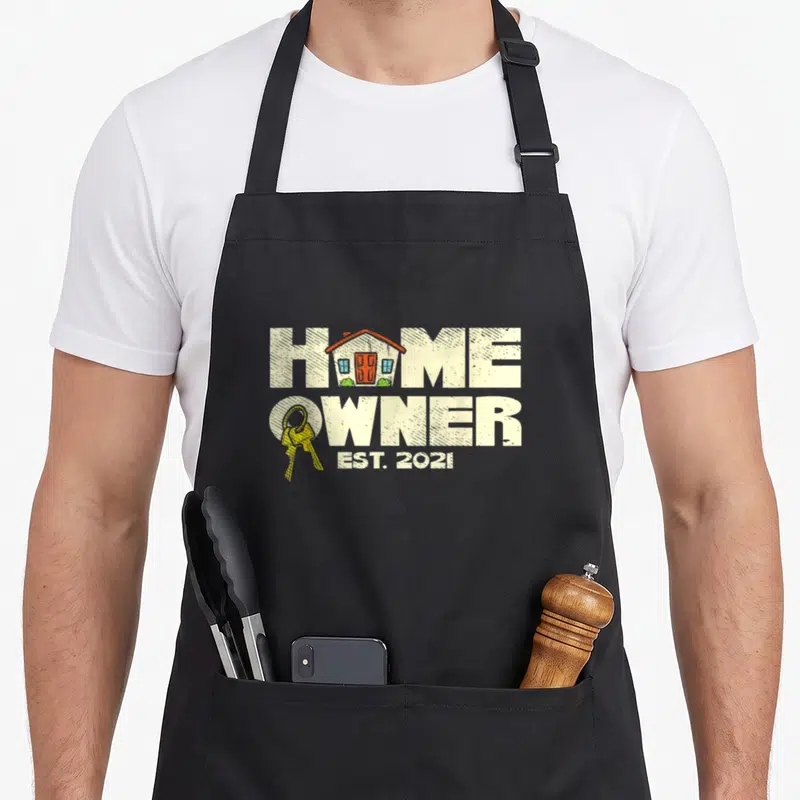 Homeowner family aprons - Distressed house graphic inside HOME text with yellow keys and EST. 2021 below — shown on a black cotton canvas apron against a white background