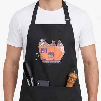 Jawa egg apron - Orange robotic hopper filled with colorful eggs, candy, and brown hooded figures in cartoon style — shown on a black cotton canvas apron against a white background
