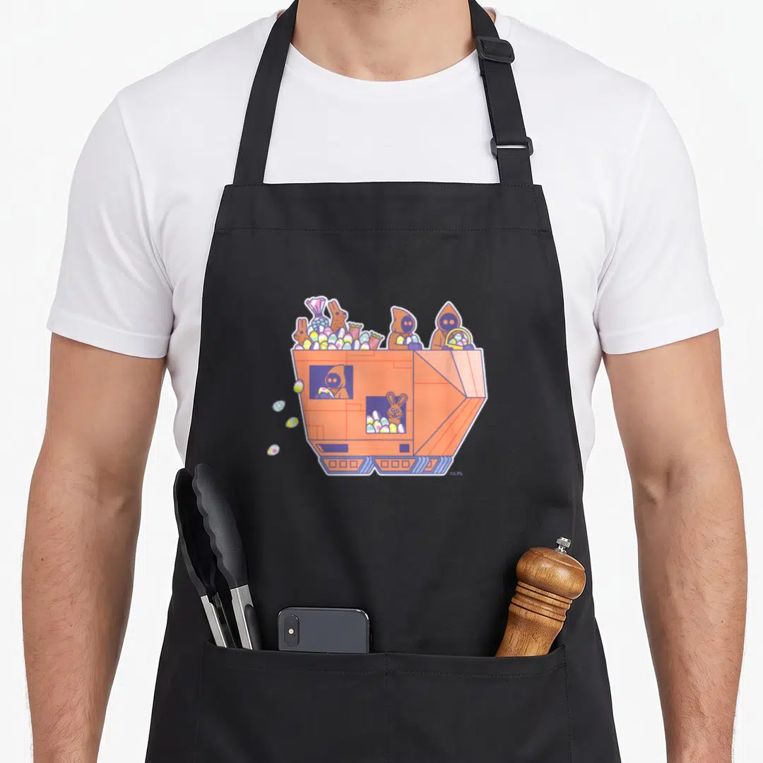 Jawa egg apron - Orange robotic hopper filled with colorful eggs, candy, and brown hooded figures in cartoon style — shown on a black cotton canvas apron against a white background