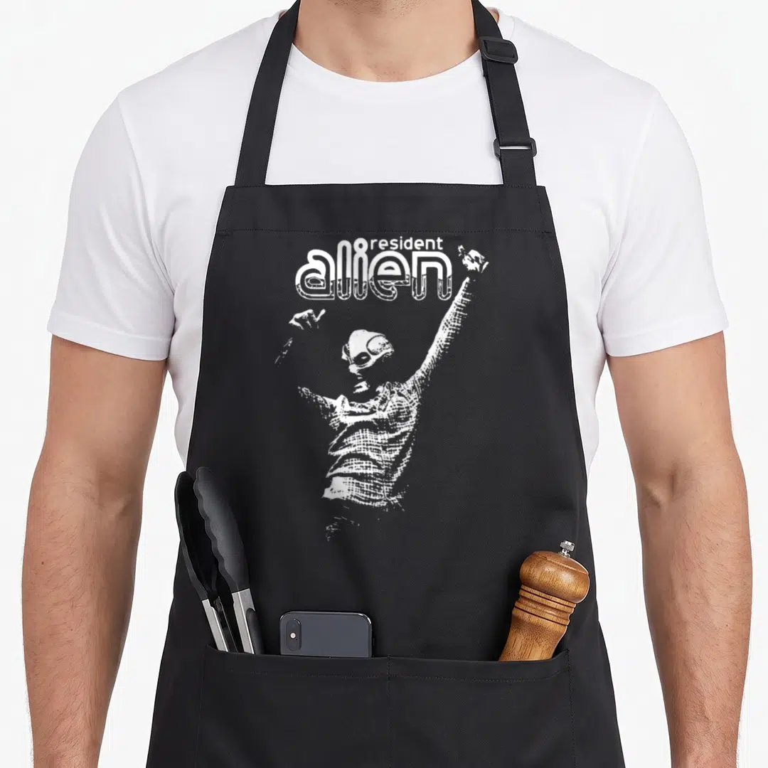 Funny alien resident apron - White line art alien figure with raised arms under resident alien text — shown on a black cotton canvas apron against a white background