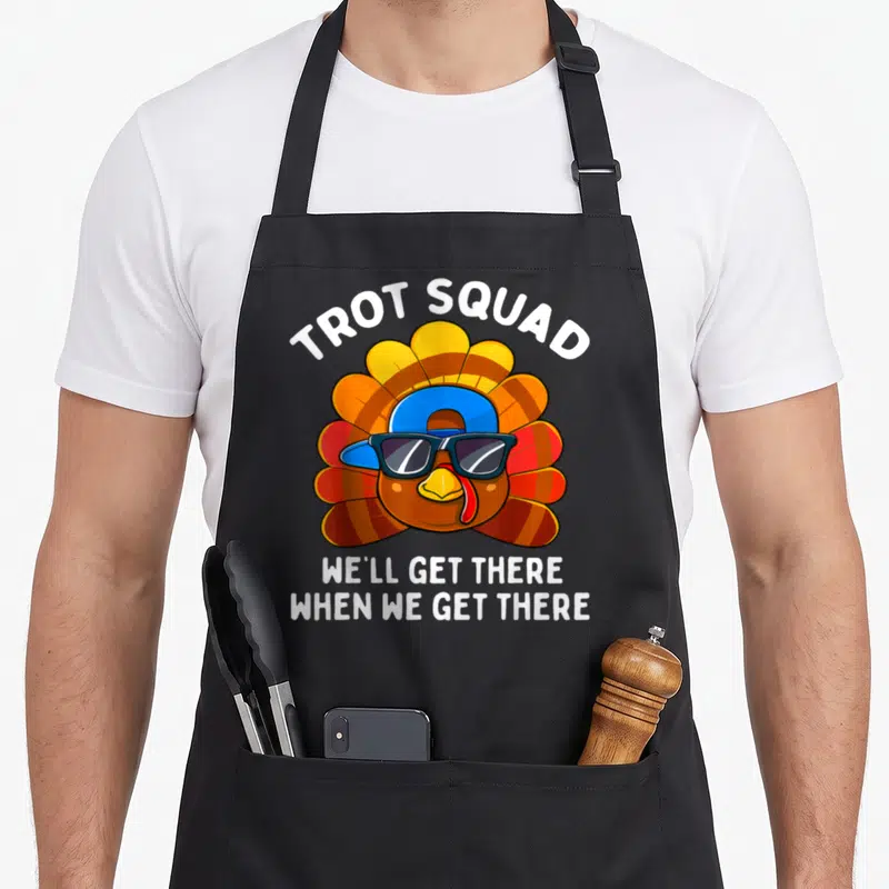 Trot thanksgiving apron - Cartoon turkey head with sunglasses and blue hat, white trot squad text and humorous quote — shown on a black cotton canvas apron against a white background