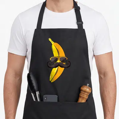 Mustache banana apron - Cartoon yellow banana wearing orange aviator sunglasses and a black handlebar mustache — shown on a black cotton canvas apron against a white background