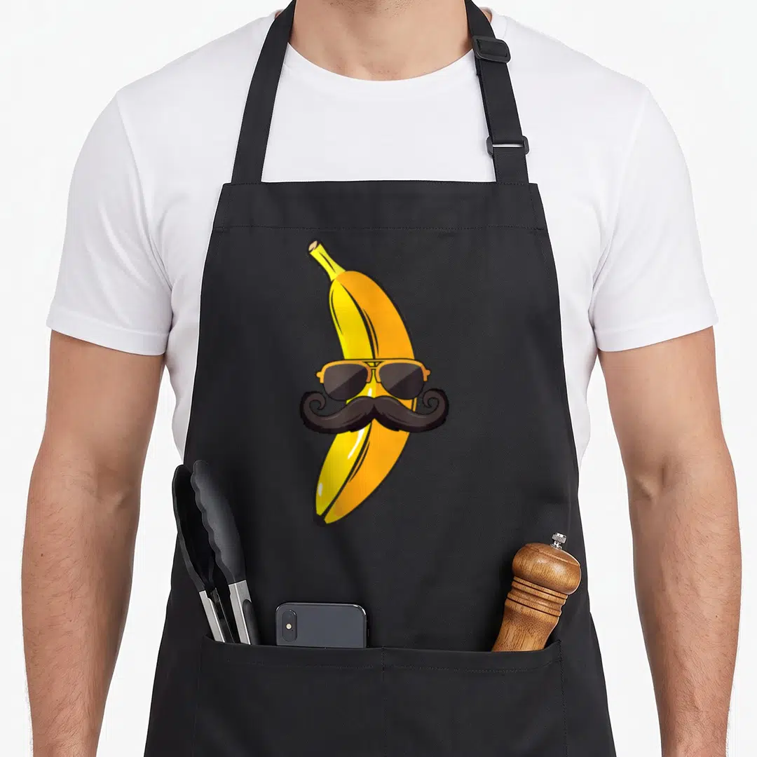 Mustache banana apron - Cartoon yellow banana wearing orange aviator sunglasses and a black handlebar mustache — shown on a black cotton canvas apron against a white background
