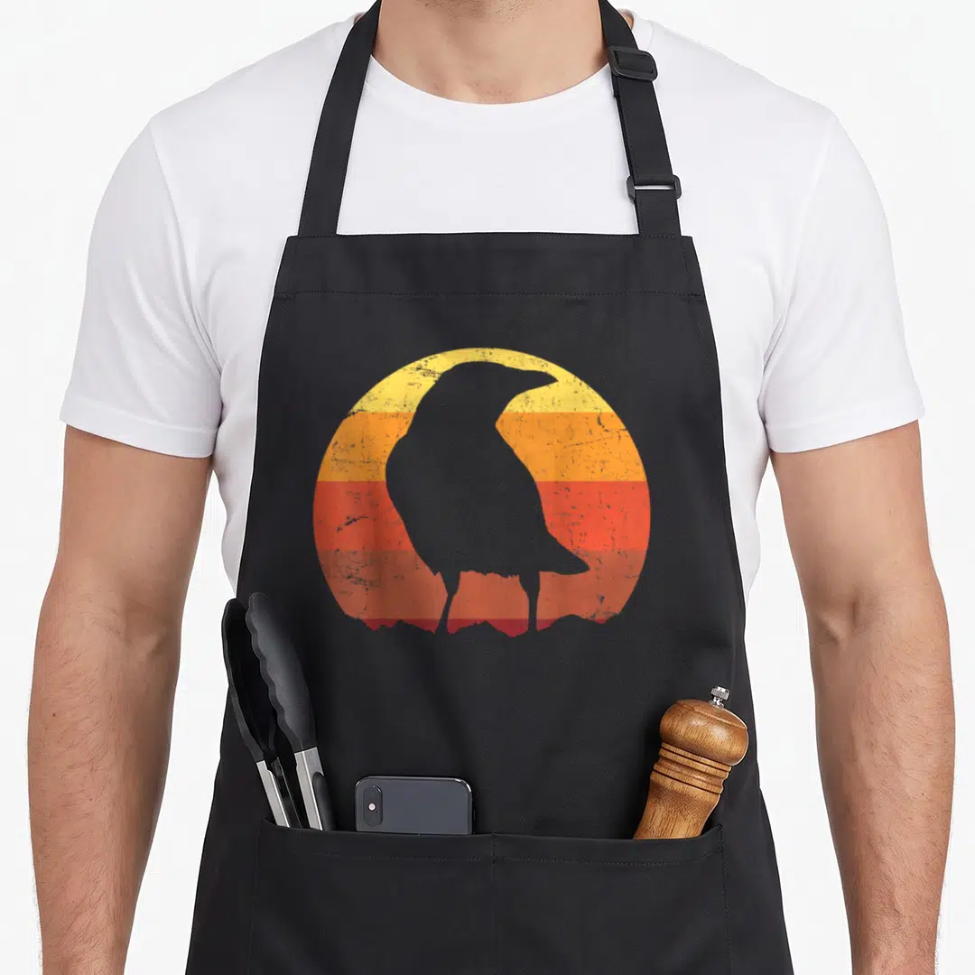 Funny crazy crow lady apron - Grey crow silhouette on striped yellow orange red retro circle — shown on a black cotton canvas apron against a white background