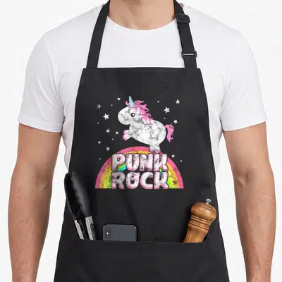 Ironic punk rock apron - Cartoon unicorn on rainbow with PUNK ROCK text and scattered stars in pink and blue — shown on a black cotton canvas apron against a white background