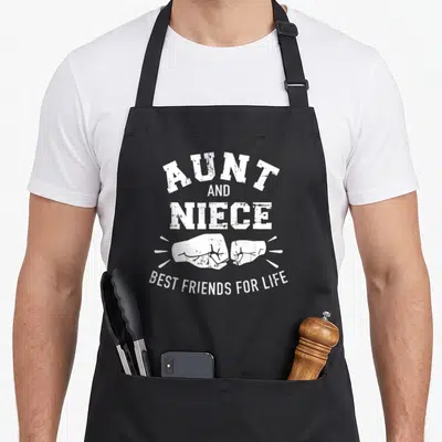 Aunt niece friends apron - White distressed fist bump graphic with stacked AUNT AND NIECE text and curved BEST FRIENDS — shown on a black cotton canvas apron against a white background