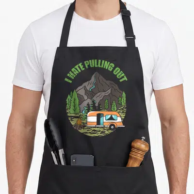 Camping travel trailer apron - I HATE PULLING OUT text over cartoon camper, mountains, trees and campfire — shown on a black cotton canvas apron against a white background