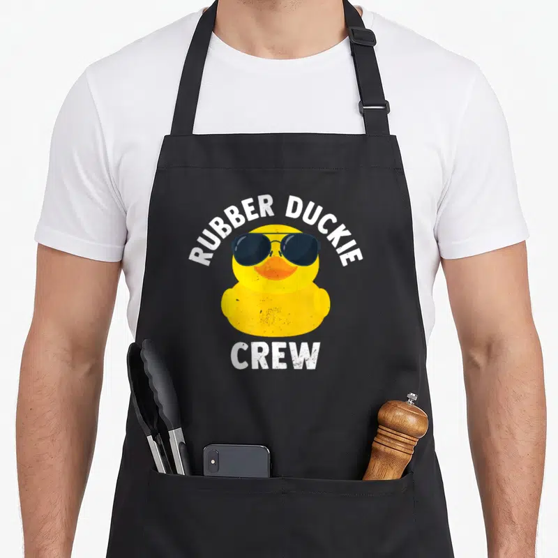 Rubber crew apron - Yellow cartoon duck wearing sunglasses with white RUBBER DUCKIE CREW text on black background — shown on a black cotton canvas apron against a white background