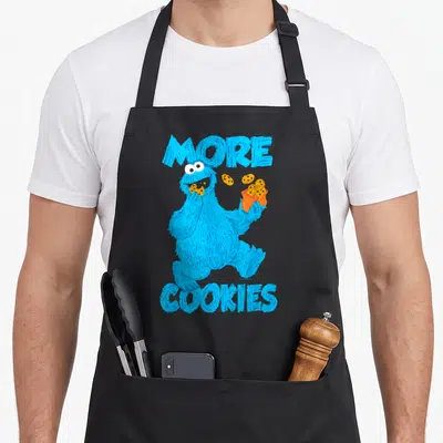 Cooky cookie monster apron - Blue character holding orange bag with floating cookies and text MORE COOKIES — shown on a black cotton canvas apron against a white background