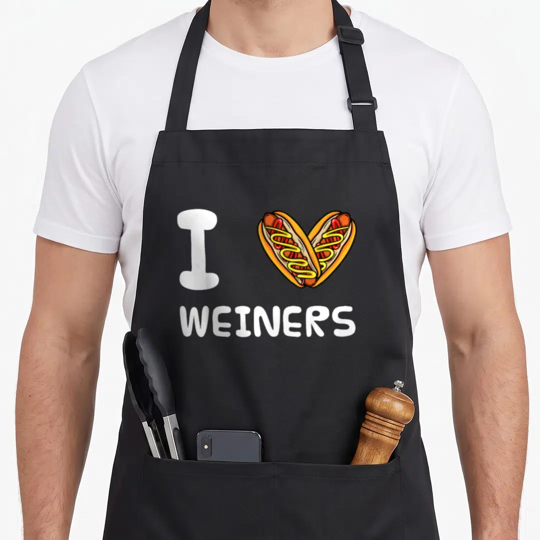 Weiner sausage apron - I WEINERS text with two hot dogs forming a heart in cartoon style — shown on a black cotton canvas apron against a white background