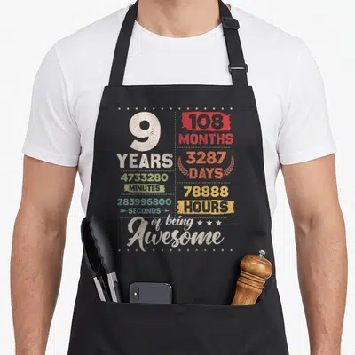 Countdown 9th birthday apron - Distressed grid of time measurements like 108 months and 3287 days surrounding a large — shown on a black cotton canvas apron against a white background