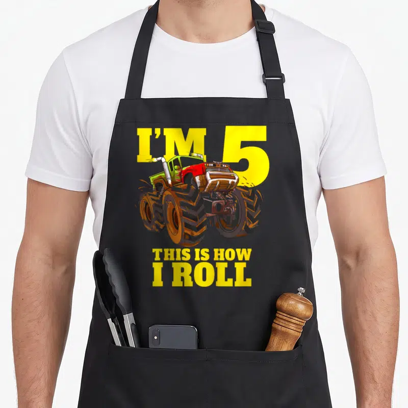 5th birthday five truck apron - Cartoon monster truck with yellow text I'm 5 This Is How I Roll on black background — shown on a black cotton canvas apron against a white background