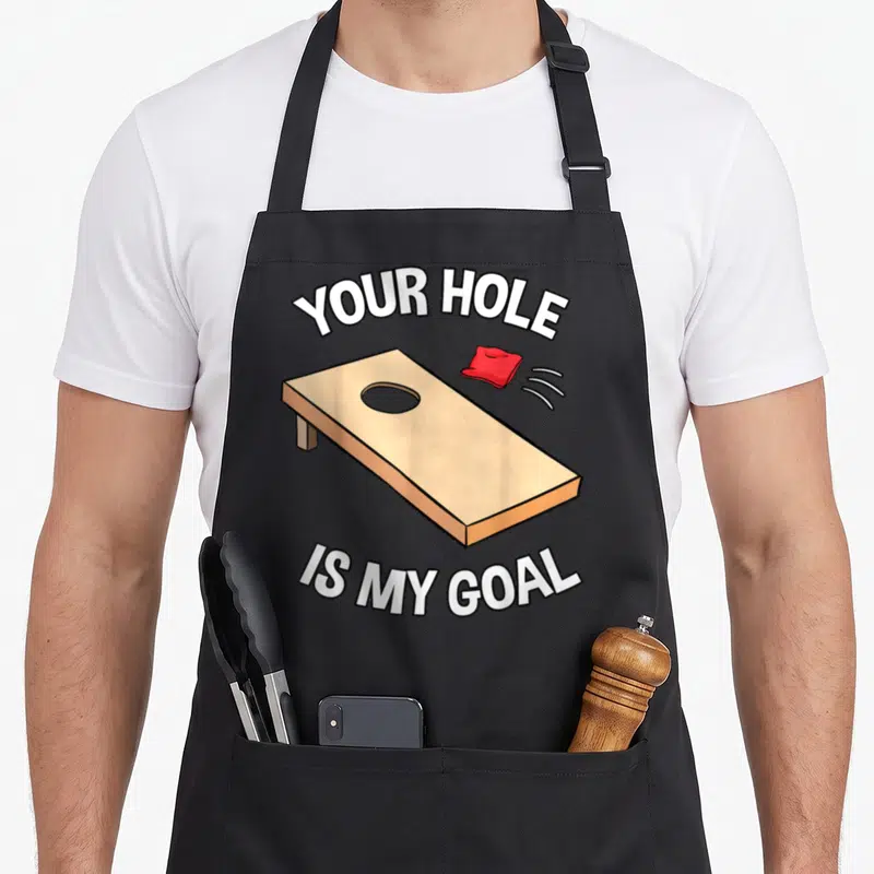 Hole bags and aprons - Tan cornhole board with red bean bag and white arched text — shown on a black cotton canvas apron against a white background