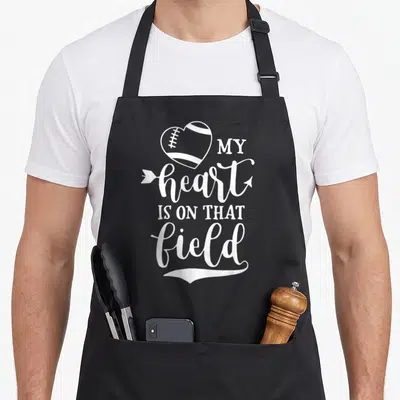 Cute field football apron - White heart shaped football and stacked text reading MY heart IS ON THAT field — shown on a black cotton canvas apron against a white background