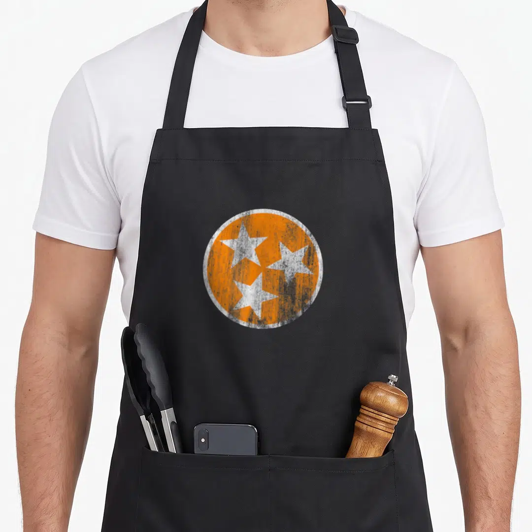 Flag volunteer apron - Distressed orange circle with three white stars in a cluster — shown on a black cotton canvas apron against a white background