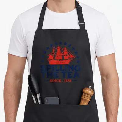 Spilling fourth of july aprons - Red ship under navy stars with Spilling The Tea Since 1773 text in retro style — shown on a black cotton canvas apron against a white background