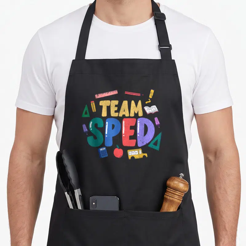 Sped cute teacher aprons - TEAM SPED text in yellow teal red blue purple with bus apple calculator ruler book triangle — shown on a black cotton canvas apron against a white background