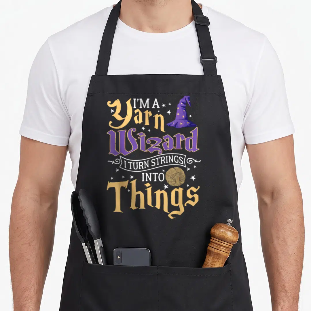 Yarn crochet apron - I'm a Yarn Wizard text with purple hat and yellow yarn ball stars — shown on a black cotton canvas apron against a white background