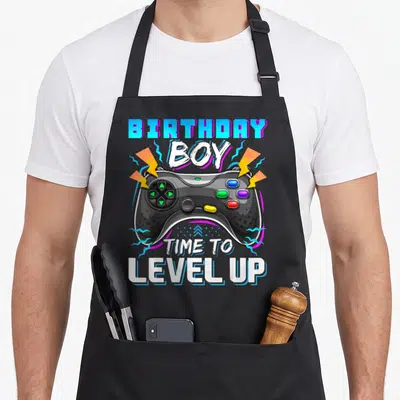 Level gamer video game apron - Game controller with birthday boy time to level up text and lightning bolts — shown on a black cotton canvas apron against a white background