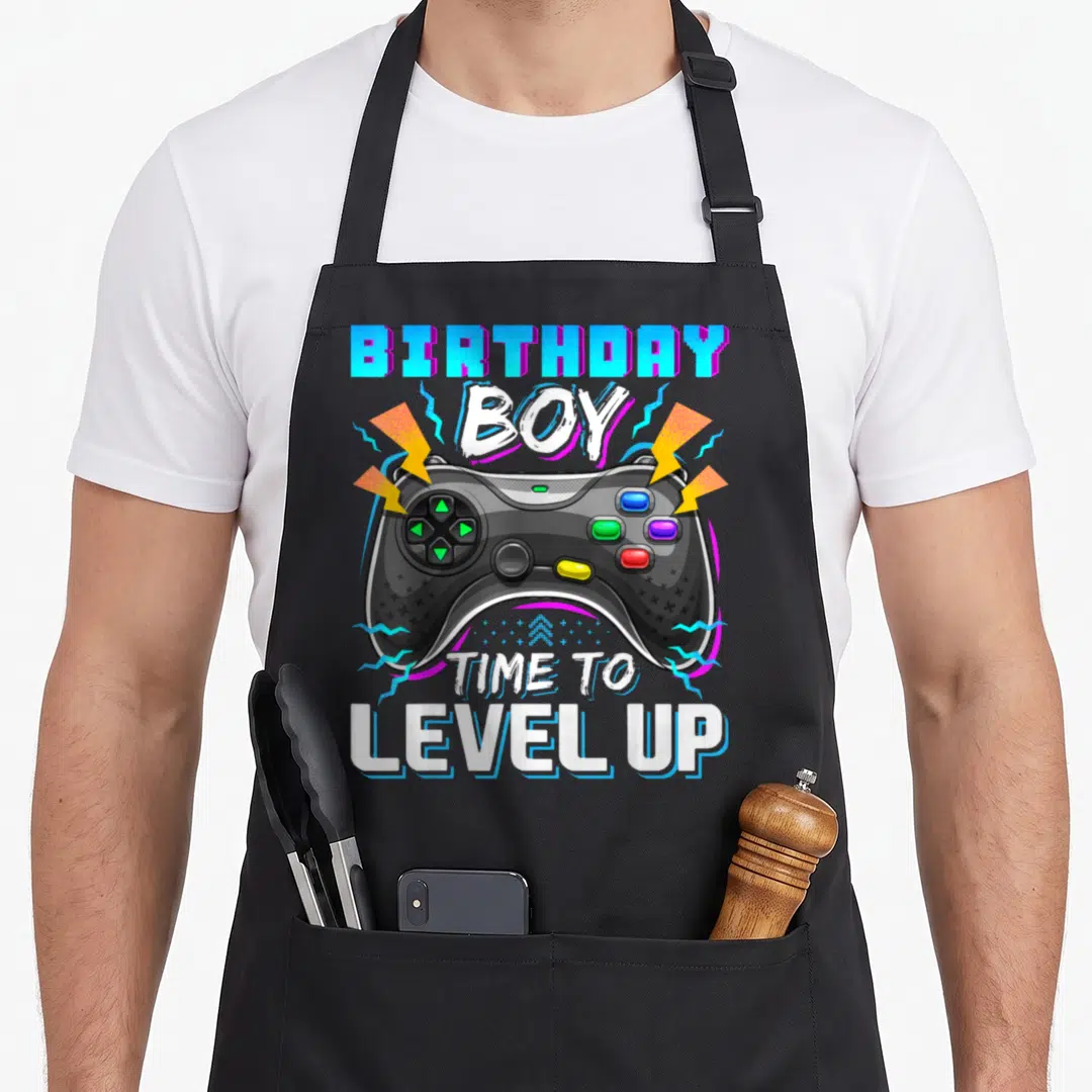Level gamer video game apron - Game controller with birthday boy time to level up text and lightning bolts — shown on a black cotton canvas apron against a white background